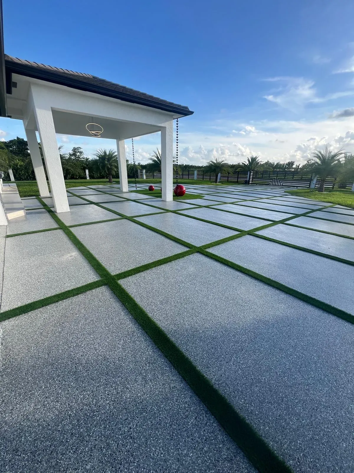Residential driveway and patio with grey flake epoxy on large concrete pads separated by strips of artificial turf, white pergola with pendant light behind