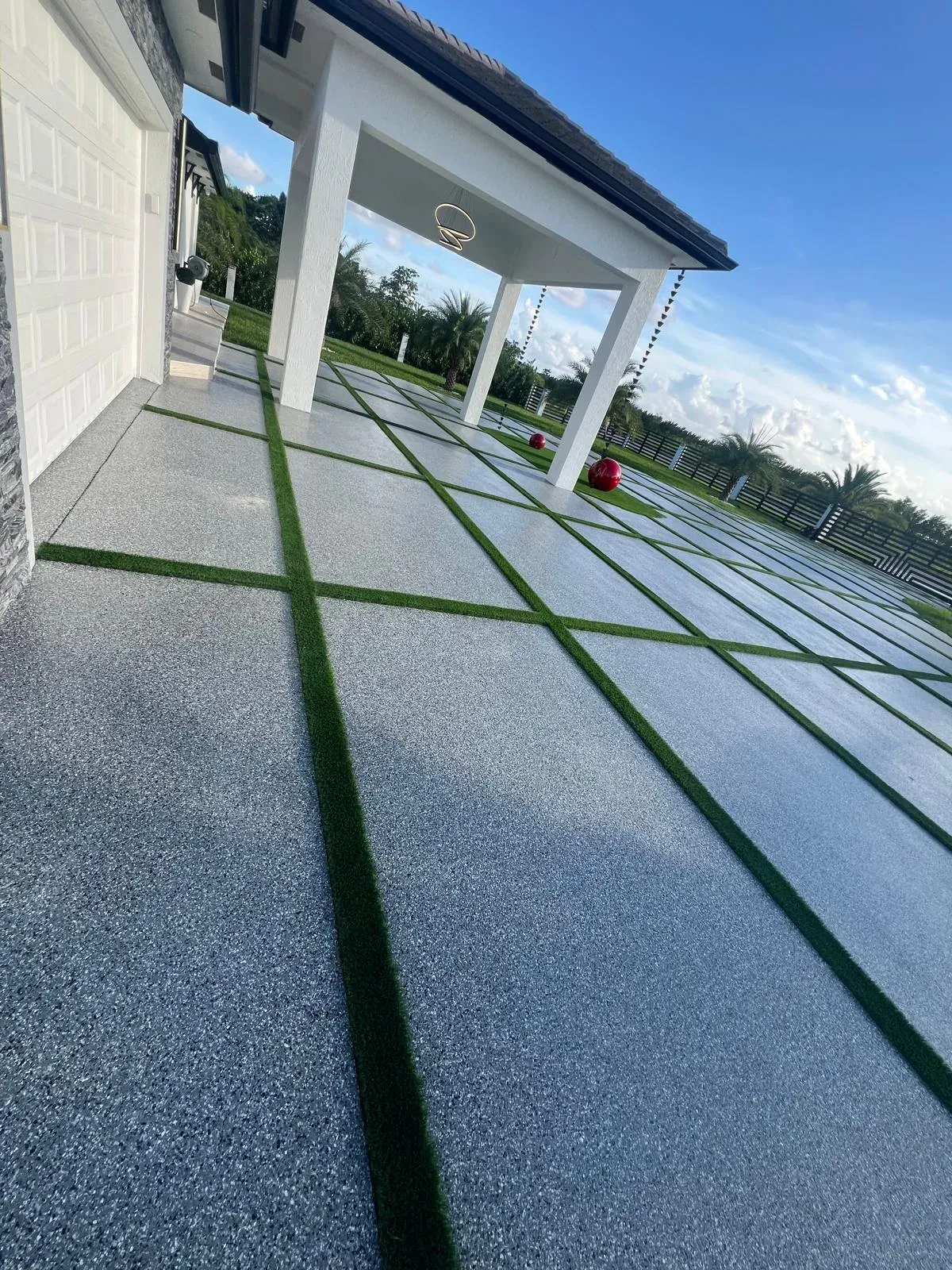 Flake epoxy driveway seen from the garage door, grey speckled pads with green turf joints running toward a covered patio