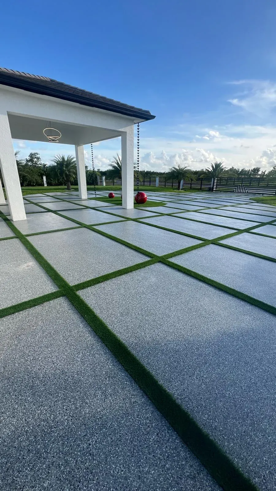 Low-angle view of a grey flake epoxy patio with turf grid lines, white pergola and palm trees under a blue sky