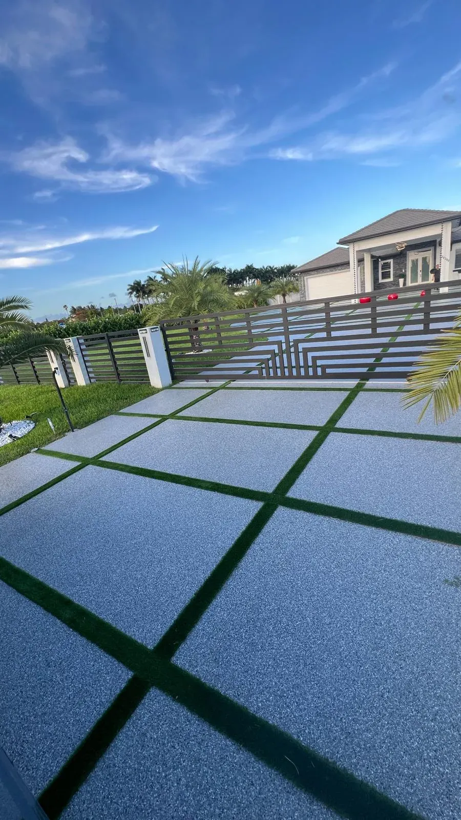 Flake epoxy driveway pads with turf joints leading to a modern slatted gate, palms and the house in the background