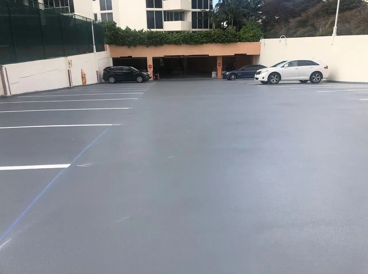 Parqueadero descubierto de un condominio con membrana de tráfico gris antideslizante y líneas blancas nuevas, con el edificio y los setos al fondo